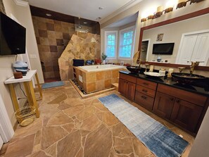 A modern bathroom with a stone accent wall, a freestanding bathtub, a large vanity with a black countertop, and a large mirror above the sink. - Heated Pool King Bed Jacuzzi Bath Arcade (Winston-Salem)
