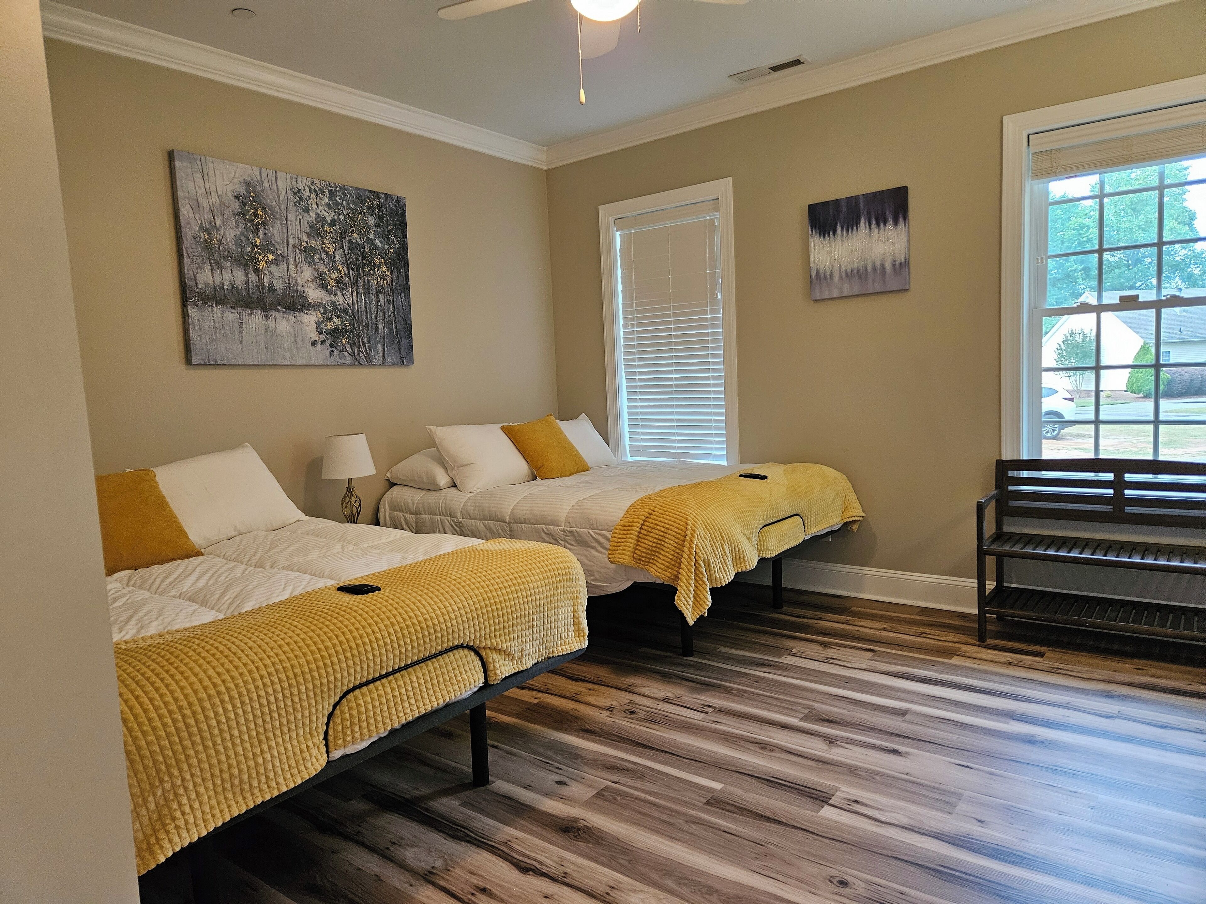 A hotel room with two beds, a window with blinds, a bench, and artwork on the walls.