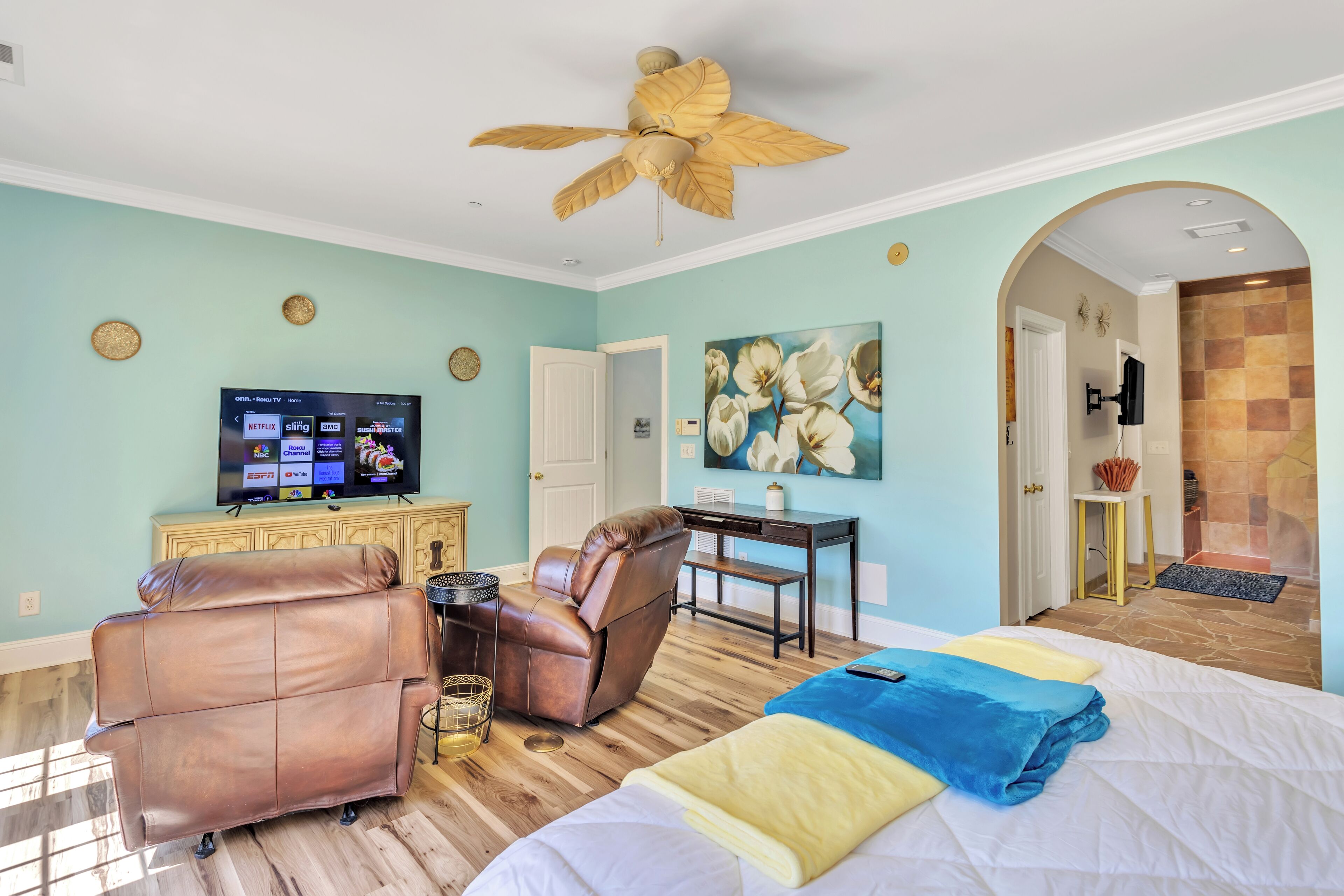 A room with a bed, a leather chair, a desk, a television, and a ceiling fan.