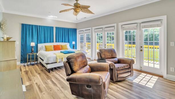 A bedroom with a bed, two leather chairs, a ceiling fan, and large windows with a view of a fenced yard. - Heated Pool King Bed Jacuzzi Bath Arcade (Winston-Salem)