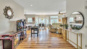 A spacious living room with a wooden floor, a sofa, a coffee table, and a foosball table. There is a kitchen in the background with a dining area. - Heated Pool King Bed Jacuzzi Bath Arcade (Winston-Salem)