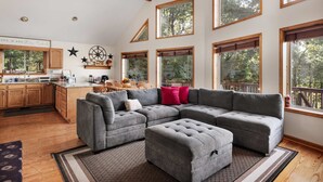 A spacious living room with a gray sectional sofa, ottoman, and a large window overlooking a deck. - Wilderness Ridge | Private Cabin w/ Mountain Views (Linden)