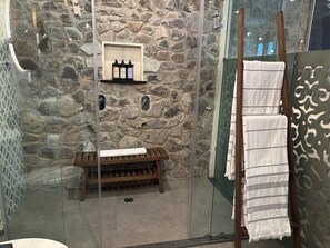 Un baño con pared de piedra, un banco de madera, estante con artículos de tocador y un perchero con toallas.