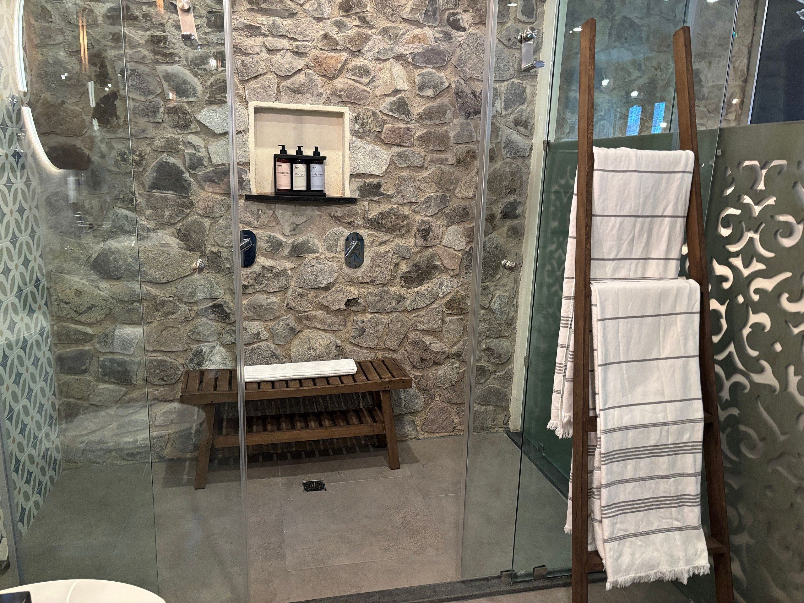A bathroom with a stone wall, a wooden bench, a shelf with toiletries, and a towel rack with towels.