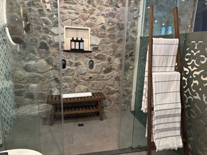 A bathroom with a stone wall, a wooden bench, a shelf with toiletries, and a towel rack with towels. - Castello di Carlo – Loft Carlo II. (Val'Quirico)