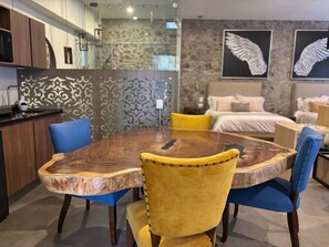 Un comedor moderno con una mesa de madera y sillas azules y amarillas, una cocina al fondo y un dormitorio con una cama y una obra de arte colgada en la pared.