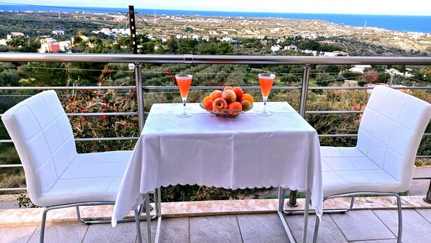 A table with a white tablecloth, two white chairs, a fruit bowl, and two drinks on a balcony overlooking a coastal landscape.