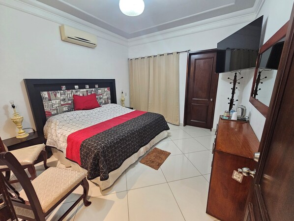 Une chambre bien rangée, avec un lit, une chaise, une table de chevet, un appareil de climatisation, une porte et un miroir.