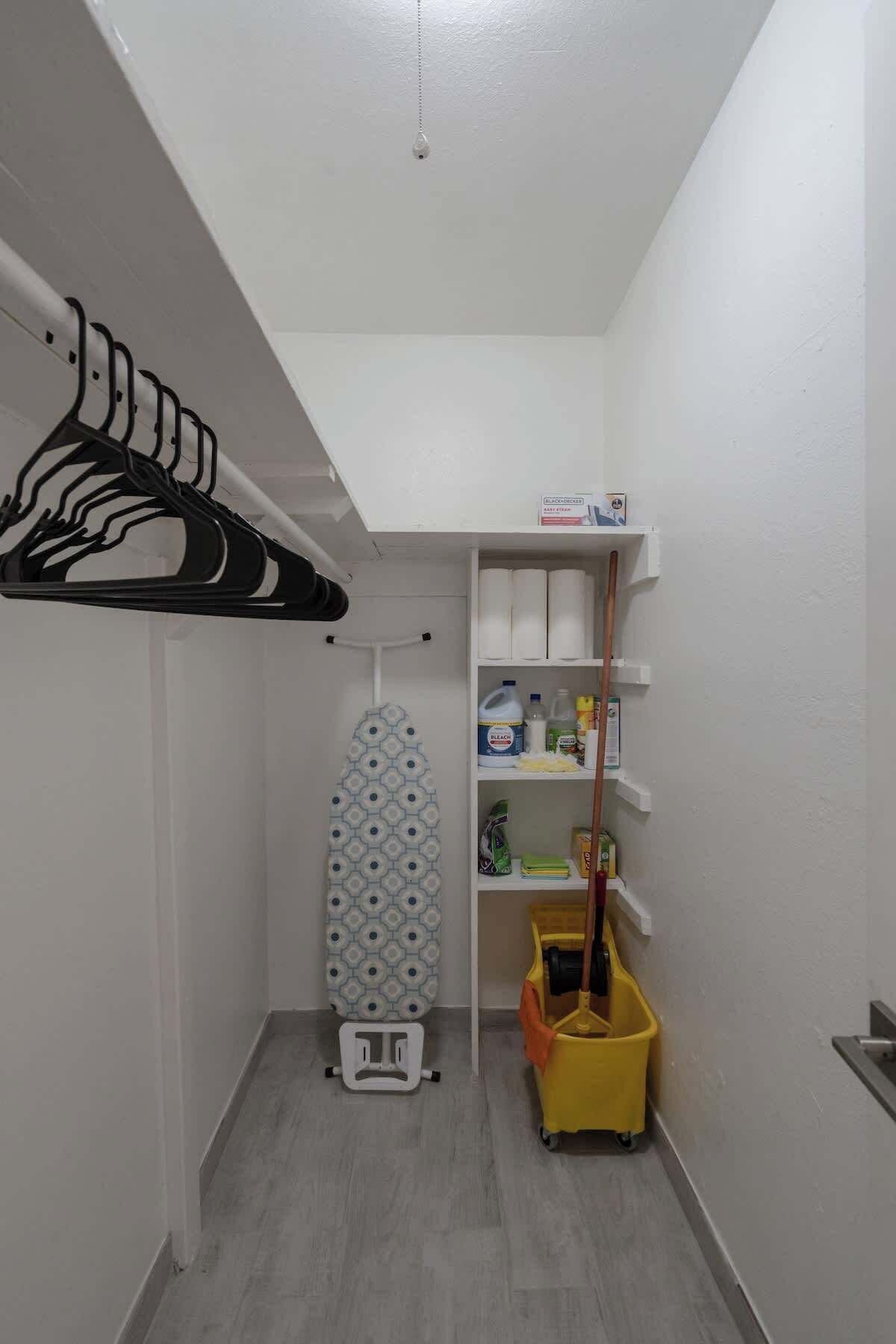 A laundry room with a drying rack, ironing board, shelves with cleaning supplies, and a mop cart.