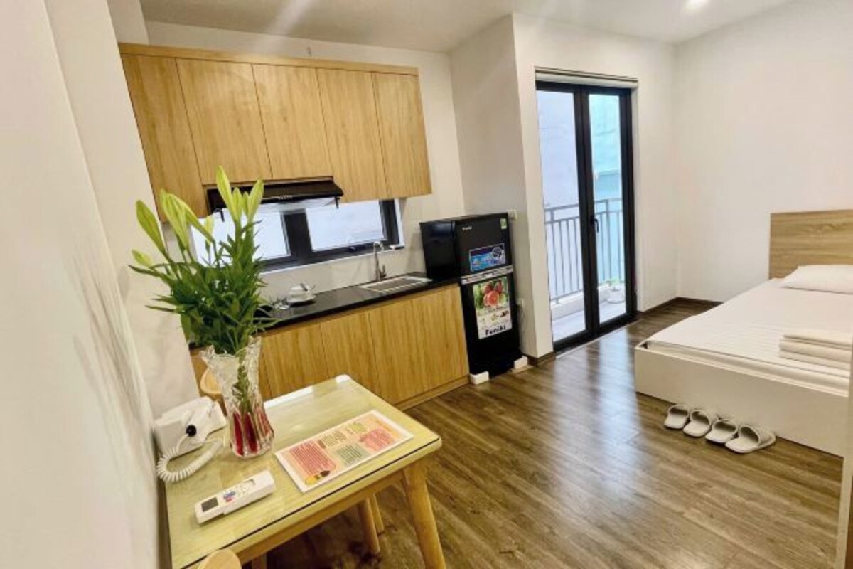 Une chambre d’hôtel compacte avec une kitchenette, une table à manger agrémentée d’une plante, un lit avec des chaussons, et une vue sur l’extérieur.