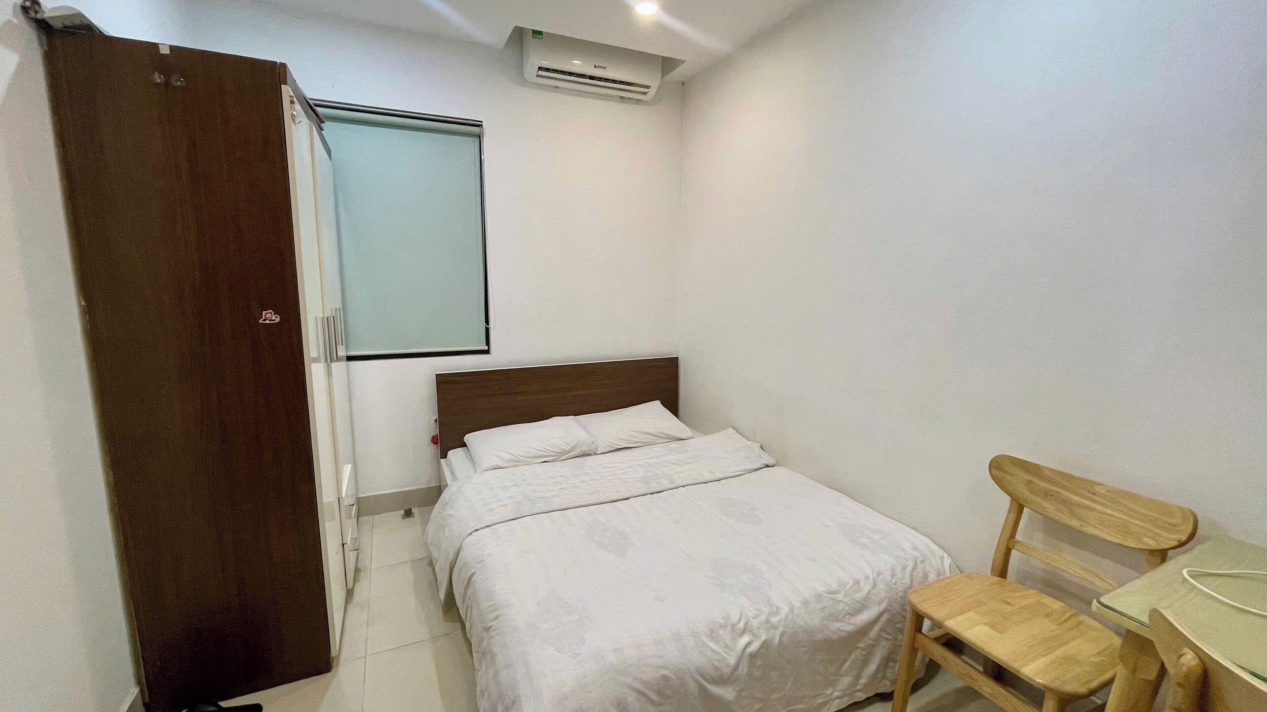 A bedroom with a bed, a wooden chair, a desk, a window with blinds, and an air conditioning unit.