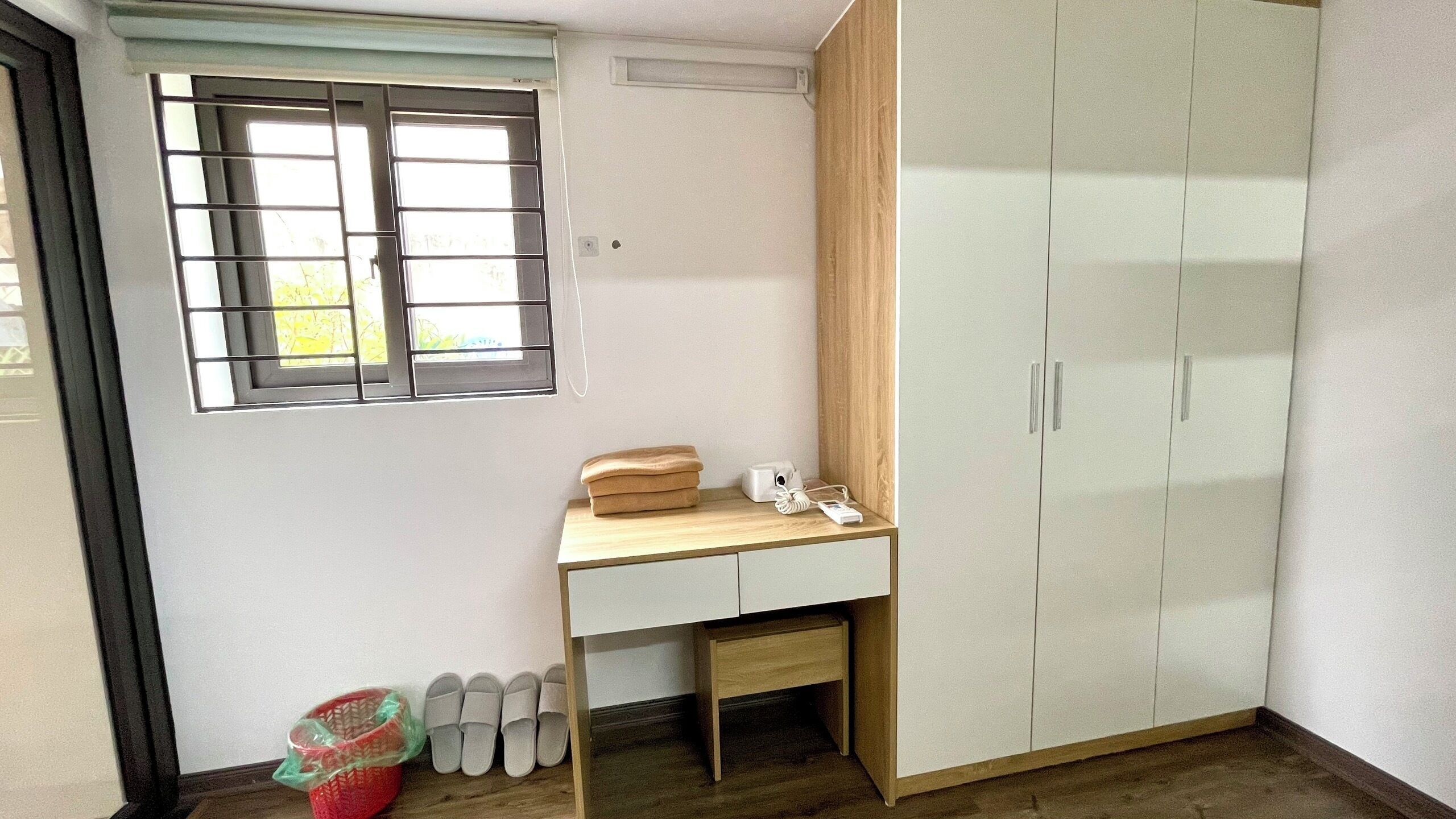 A room with a window, a desk, a wardrobe, and a trash bin.