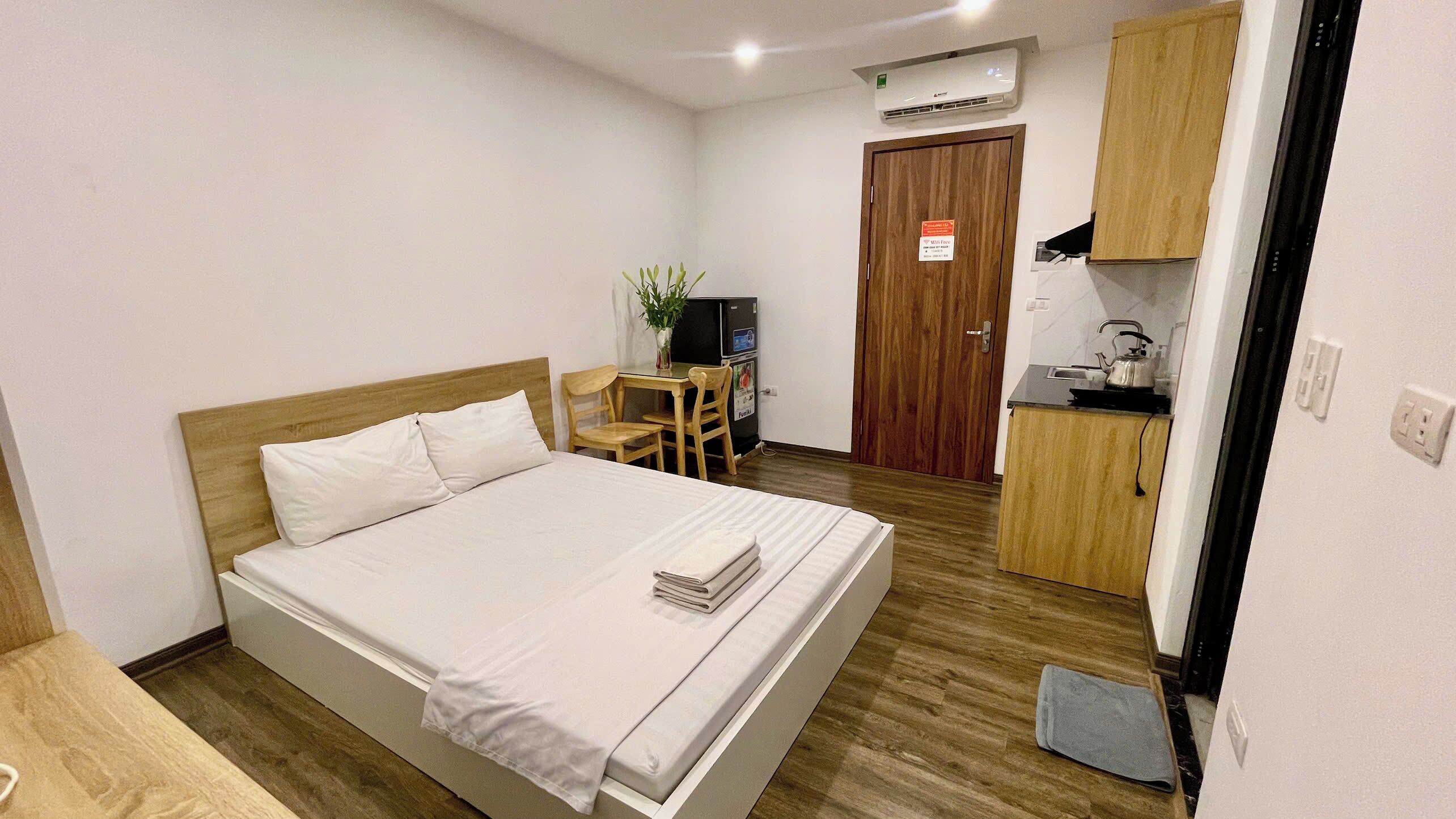 A hotel room with a bed, a small desk with a chair, a kitchenette, and a door.