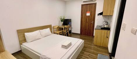 A hotel room with a bed, a small desk with a chair, a kitchenette, and a door.