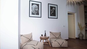 A room with two upholstered chairs, a small wooden table with a vase and incense sticks, and two framed black-and-white photographs on the wall.