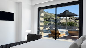 A modern hotel room with a large bed, a view of a pool area with lounge chairs and umbrellas, and a scenic mountain backdrop. - Villa Christy, Villa Christy (Mykonos)