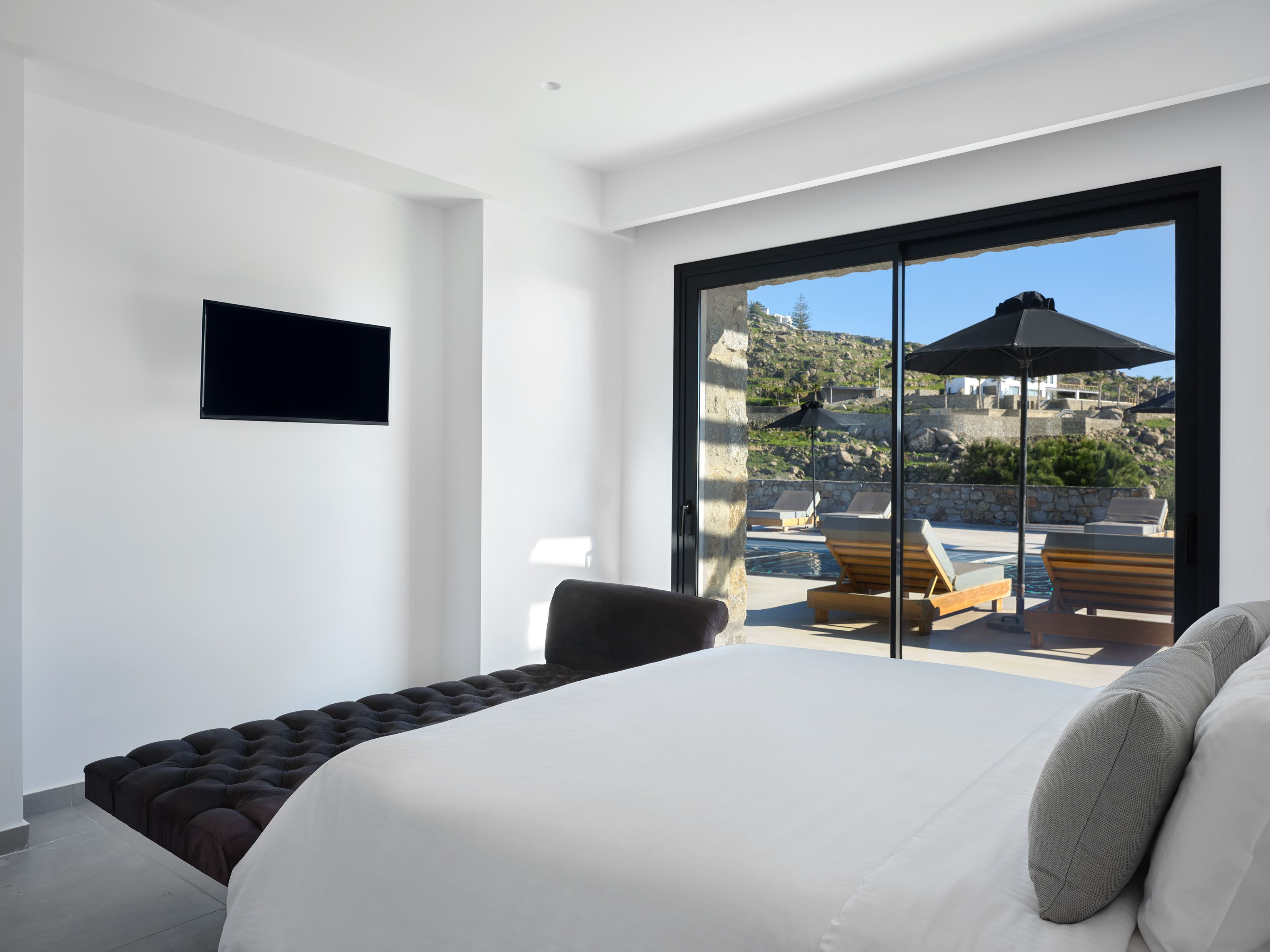 A modern bedroom with a large bed, a TV mounted on the wall, and a view of a pool and outdoor seating area.