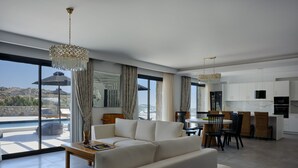 A modern living room with a sofa, dining table, and chairs, a kitchen area, and large windows with a view of the landscape. - Villa Christy, Villa Christy (Mykonos)