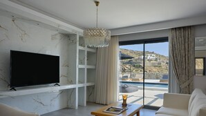 A modern living room with a flat-screen TV, a wooden coffee table, and a sofa. There is a large window with a view of a pool and a mountainous landscape. - Villa Christy, Villa Christy (Mykonos)