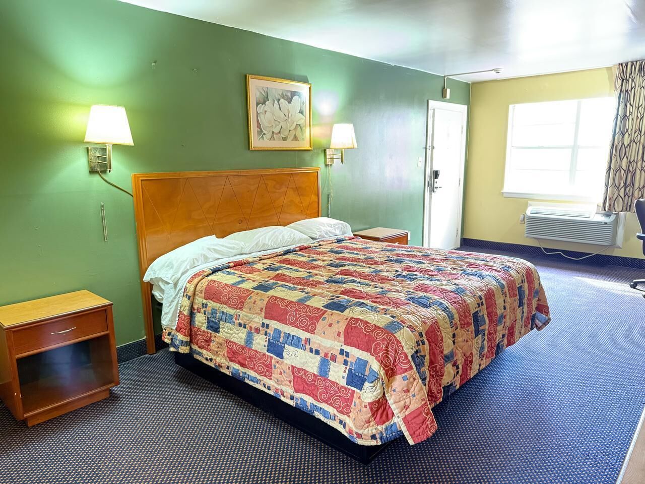 A hotel room with a bed, two bedside lamps, a nightstand, a window with curtains, and a wall-mounted picture.