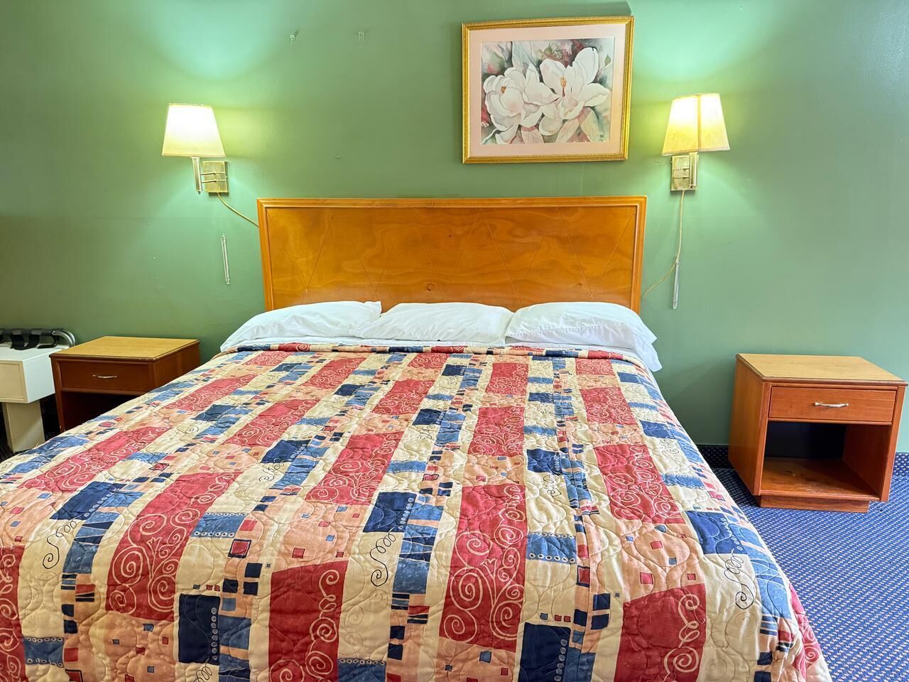 Une chambre d’hôtel avec une tête de lit en bois, une couvre-lit matelassée, deux lampes de chevet, une table de chevet et une petite armoire en bois.