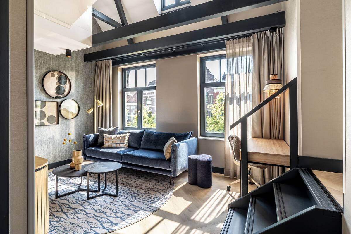 Ein modernes Wohnzimmer mit einem blauen Sofa, einem runden Couchtisch und einem gemusterten Teppich. Zwei Fenster mit Vorhängen, eine Treppe und eine Wandleuchte sind ebenfalls zu sehen.