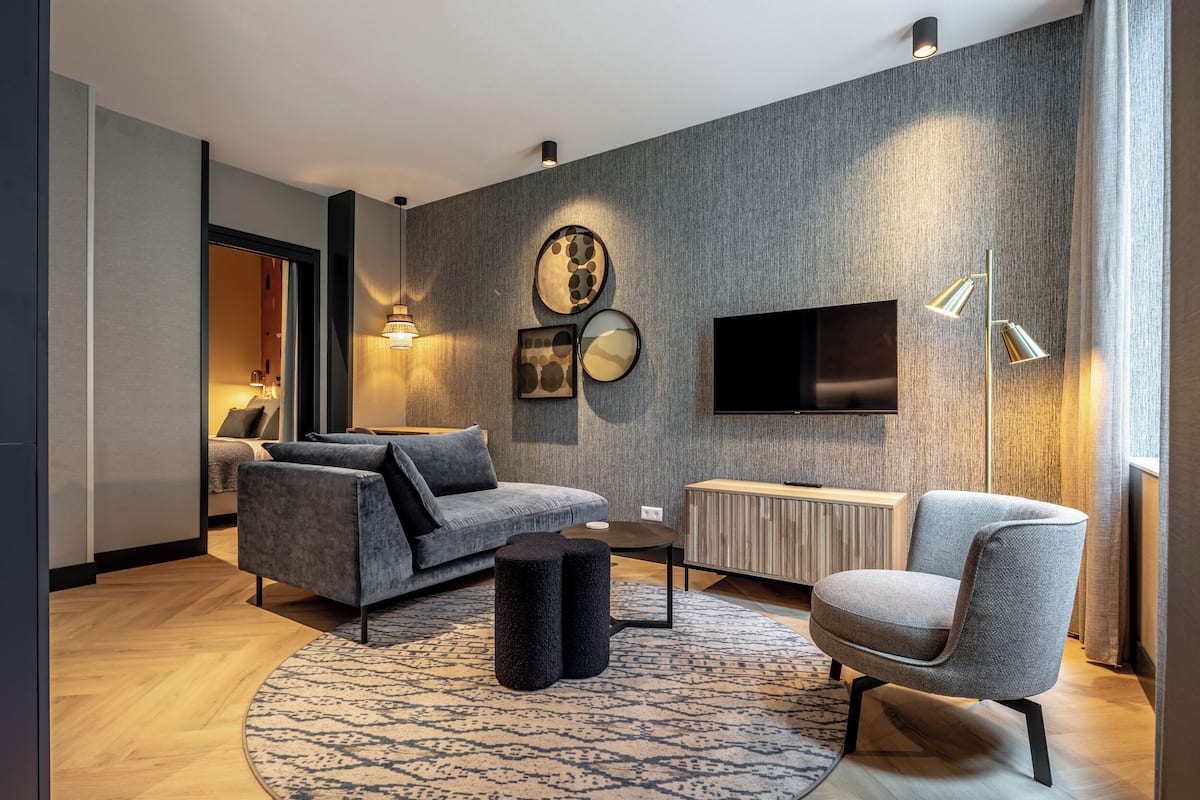 Ein modernes Wohnzimmer mit einem grauen Sofa, einem runden Couchtisch und einem grauen Sessel. An der Wand ist ein Flachbildfernseher angebracht und es steht eine Stehlampe im Raum.