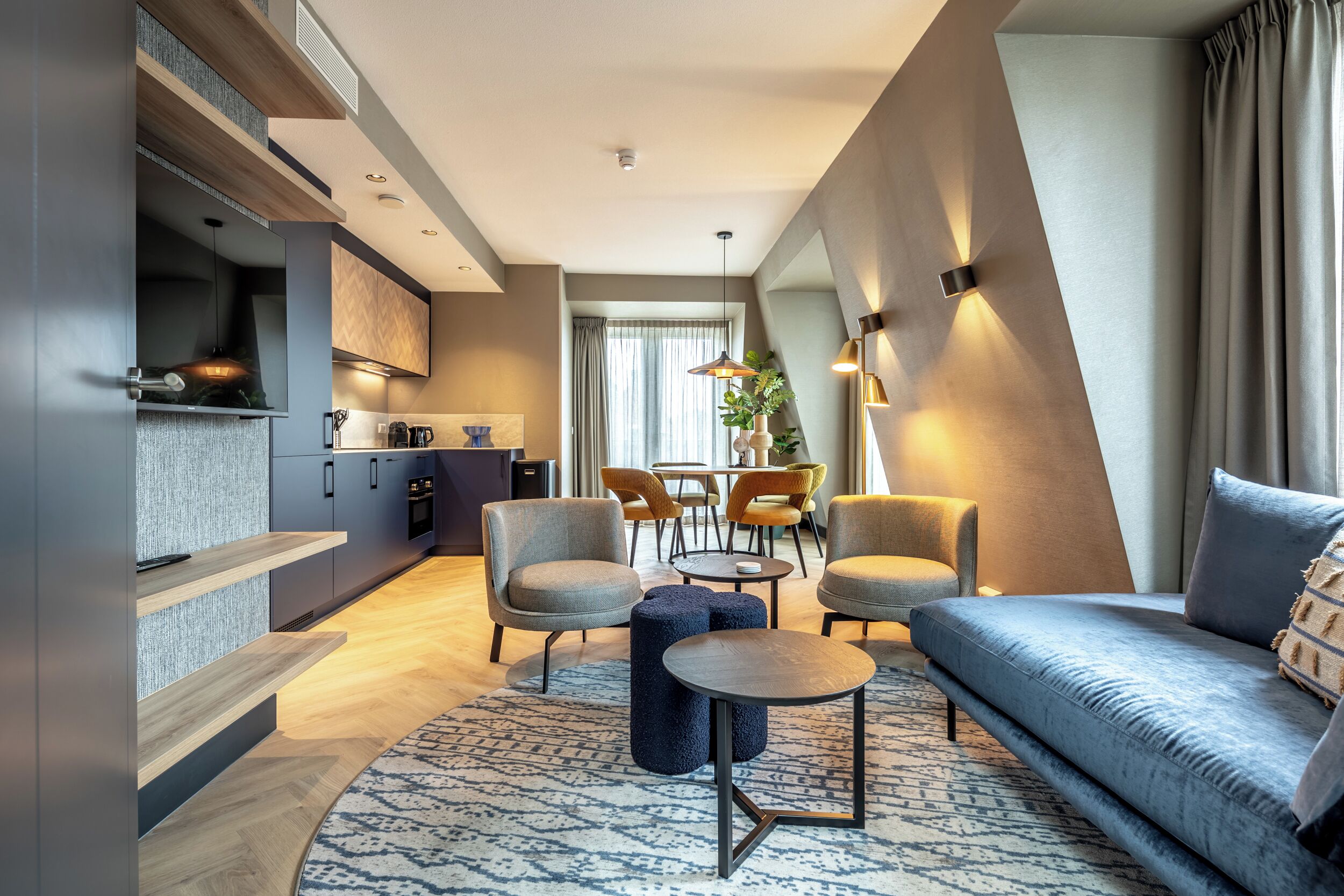 A modern living room with a sofa, armchairs, and a coffee table. There is a kitchen area with cabinets and a dining table with chairs.