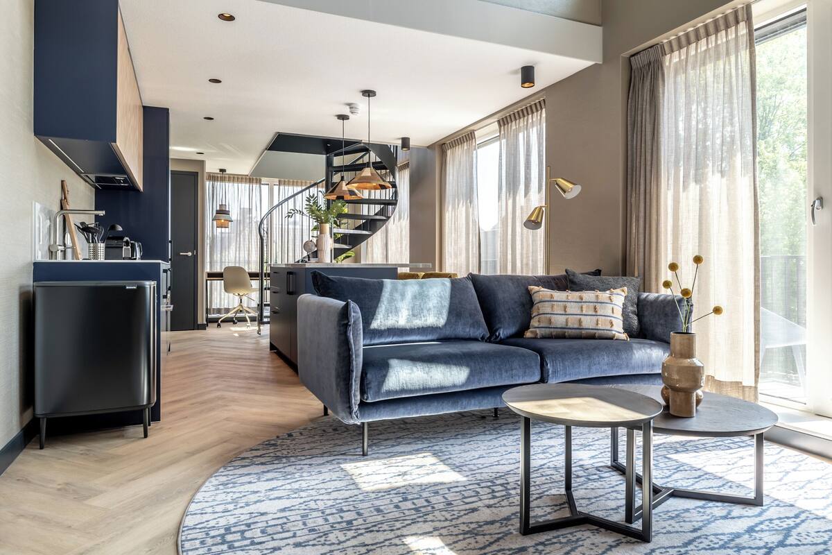 Ein modernes Wohnzimmer mit einem blauen Sofa, einem runden Couchtisch und einer Wendeltreppe im Hintergrund.