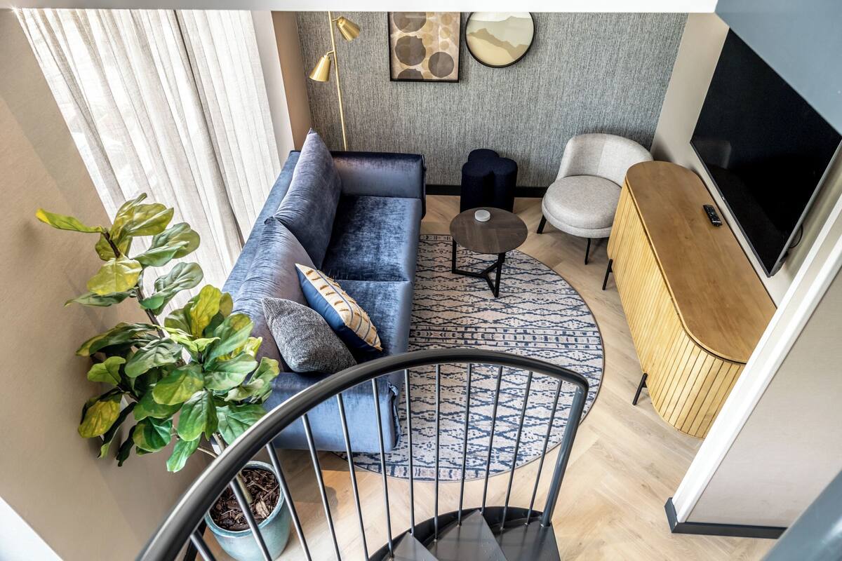 Ein modernes Wohnzimmer mit einem blauen Sofa, einem runden Couchtisch, einem grauen Teppich mit Muster, einer Holzkommode und einem Fernseher.