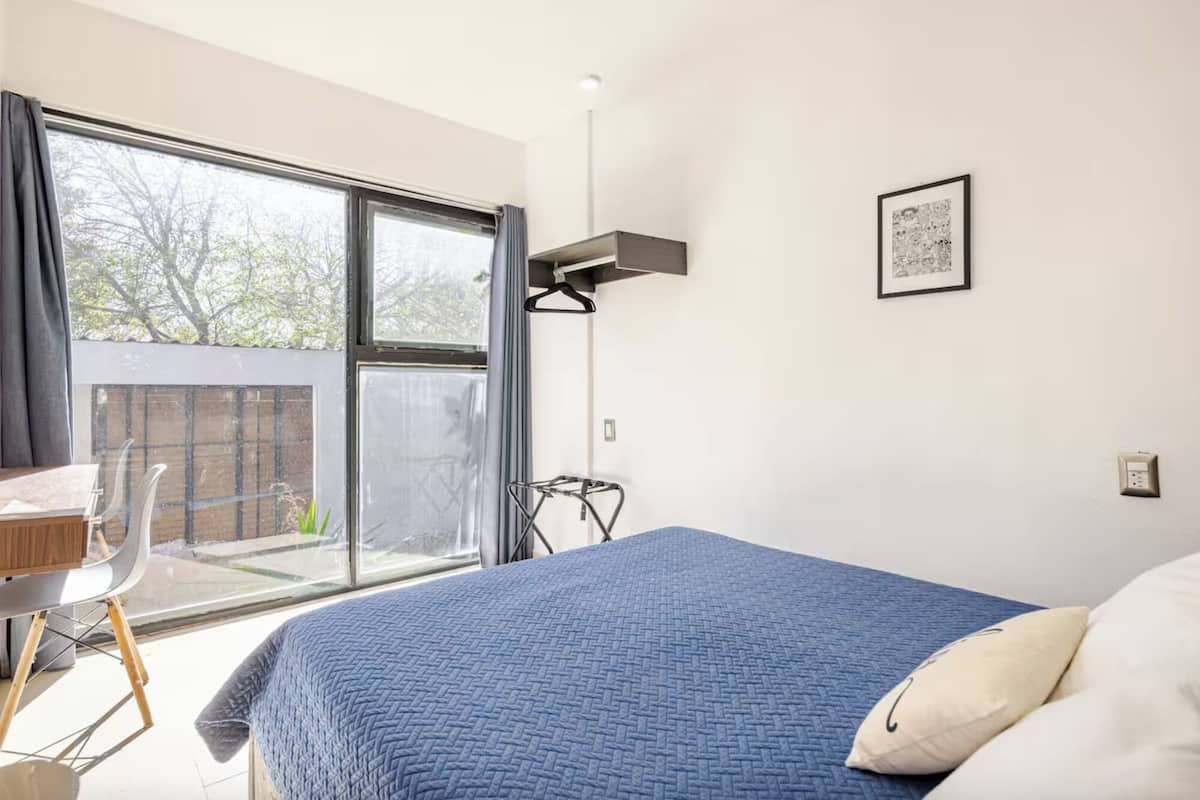 Un dormitorio con una cama grande, un escritorio y una silla. Hay una ventana con cortinas, un cuadro en la pared y una lámpara colgante.