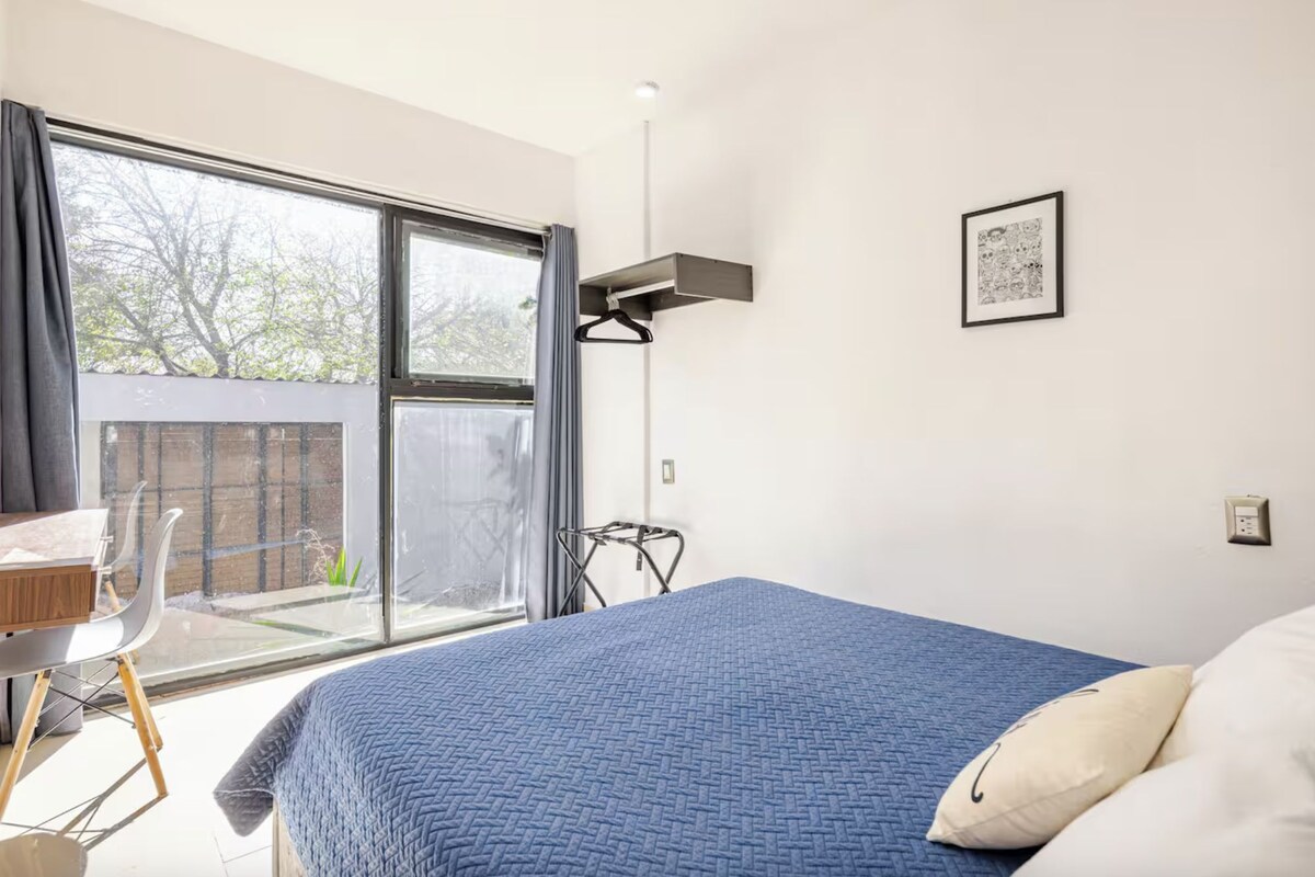 A bedroom with a large bed, a desk, and a chair. There is a window with curtains, a picture on the wall, and a hanging light fixture.