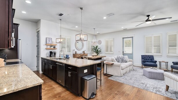 A modern kitchen with granite countertops, a central island, and dark cabinetry. There is a dining area with a white sofa and blue chairs. The room has wooden flooring and white walls with window treatments.