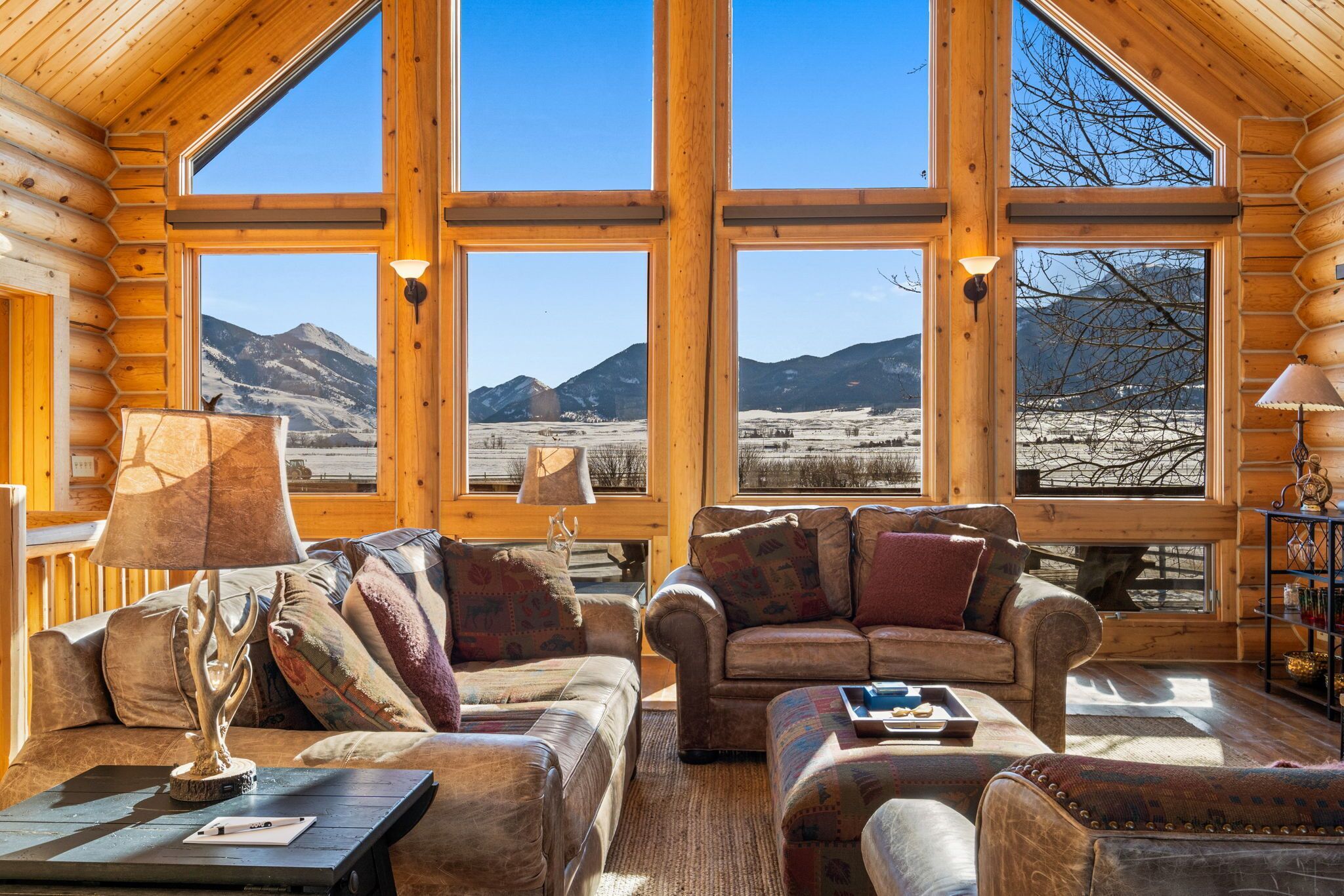 L’intérieur d’une cabane en rondins, avec de grandes fenêtres, une cheminée et un coin salon confortable.