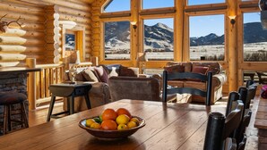 Innenansicht einer Holzblockhütte mit Essbereich, Kamin und großen Fenstern mit Blick auf die Berge.