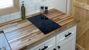 Een keuken met houten planken met witte serviesdelen en glazen, een zwart inductiekookveld en een raam met zonwering.