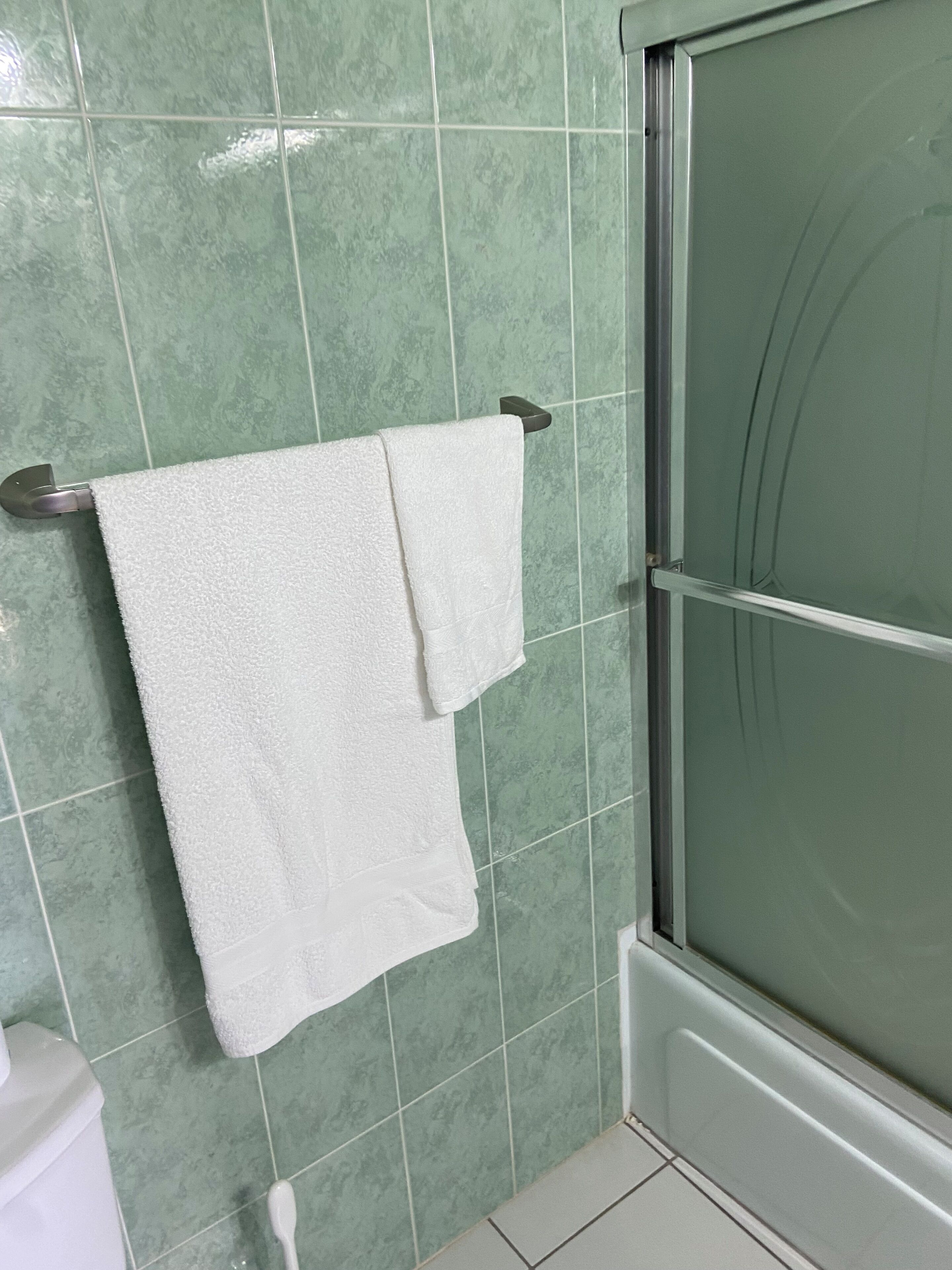 A bathroom with green tiles, a white towel hanging on a metal towel rack, and a glass shower door.