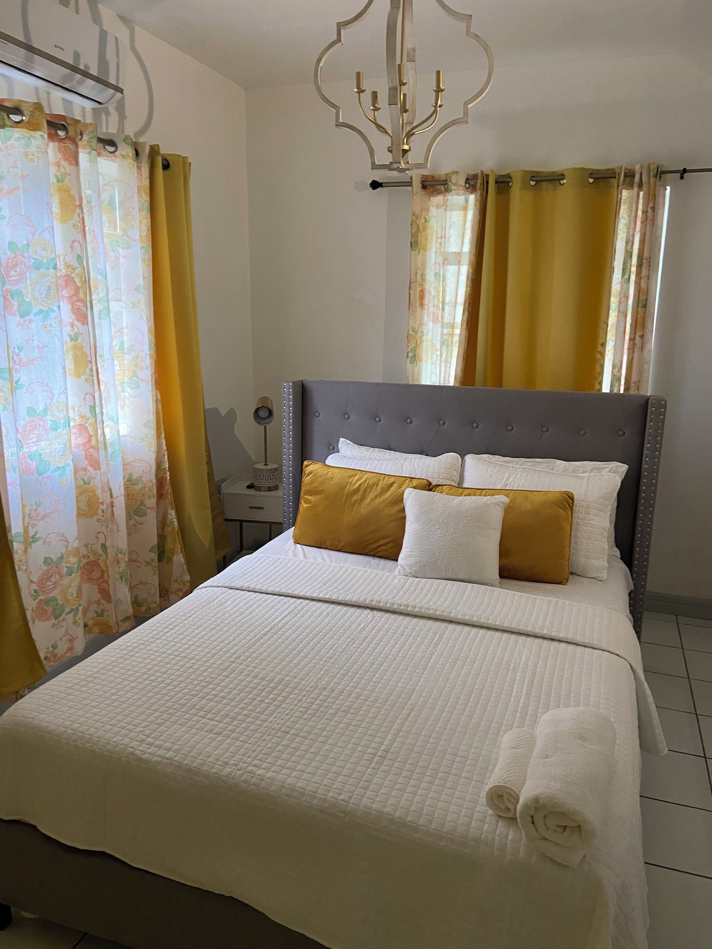 A neatly made bed with a white quilted bedspread and two pillows, one white and one mustard yellow, in a room with a grey upholstered headboard and a chandelier.