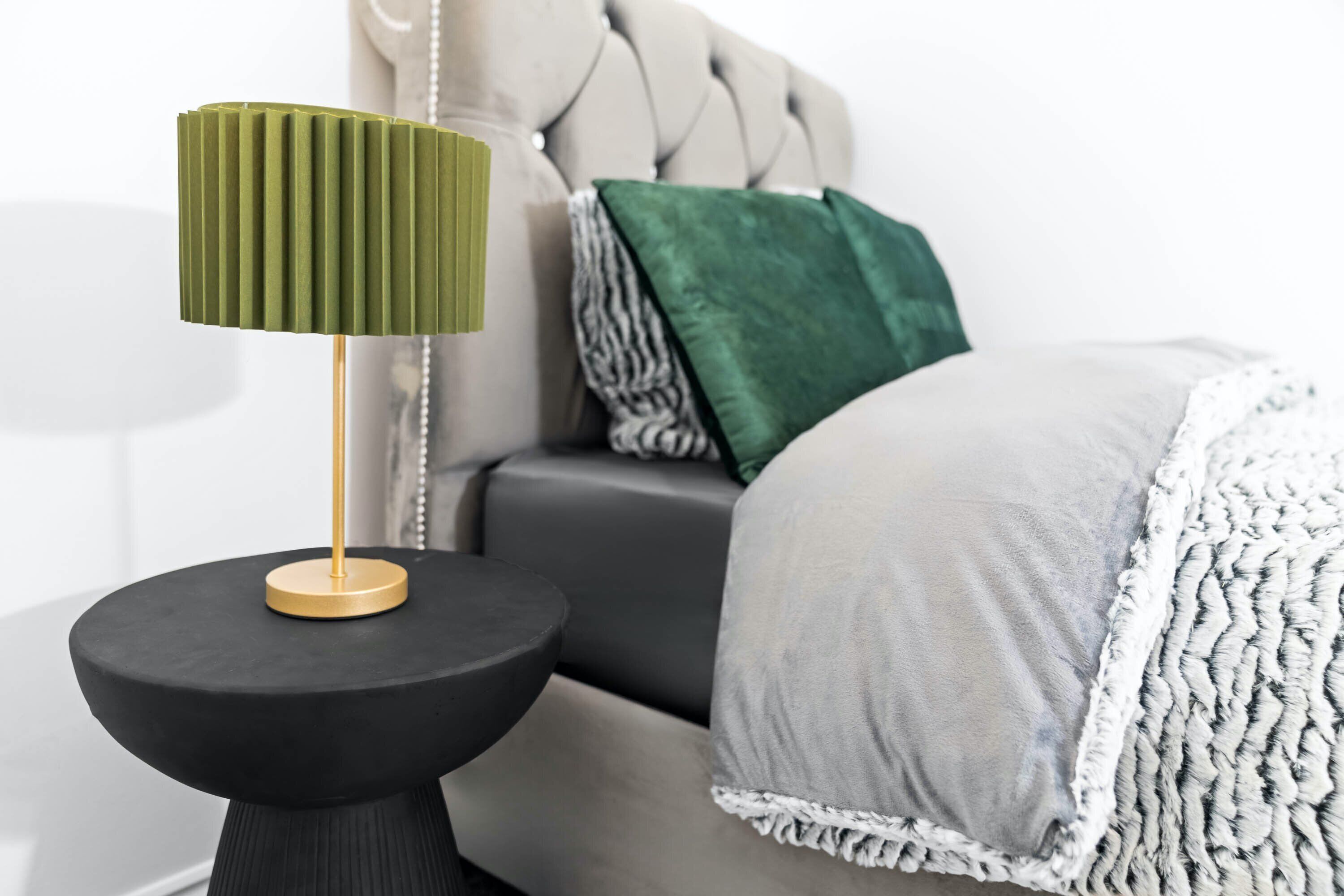 A modern bedroom with a tufted headboard, a round black side table with a green lamp, and a grey sofa with patterned cushions.