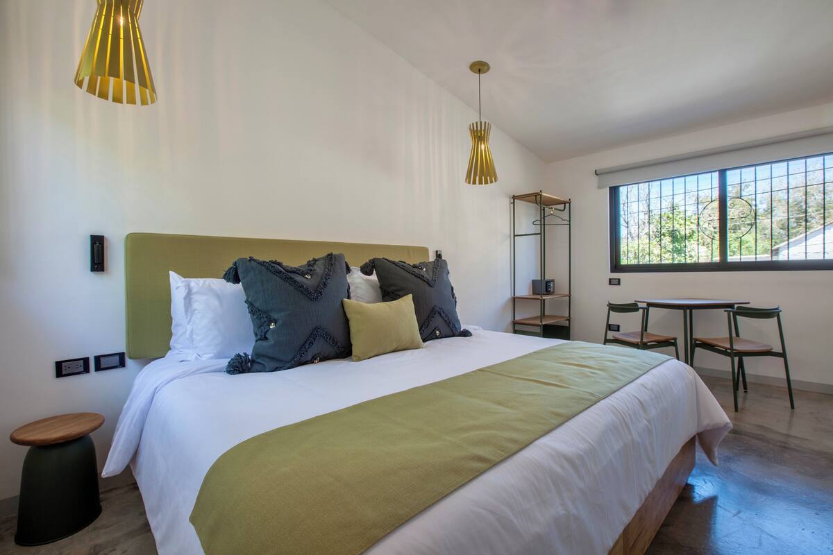 Una habitación de hotel moderna con una cama grande, dos lámparas colgantes, una pequeña mesa con sillas y un ventanal con vistas a zonas verdes.