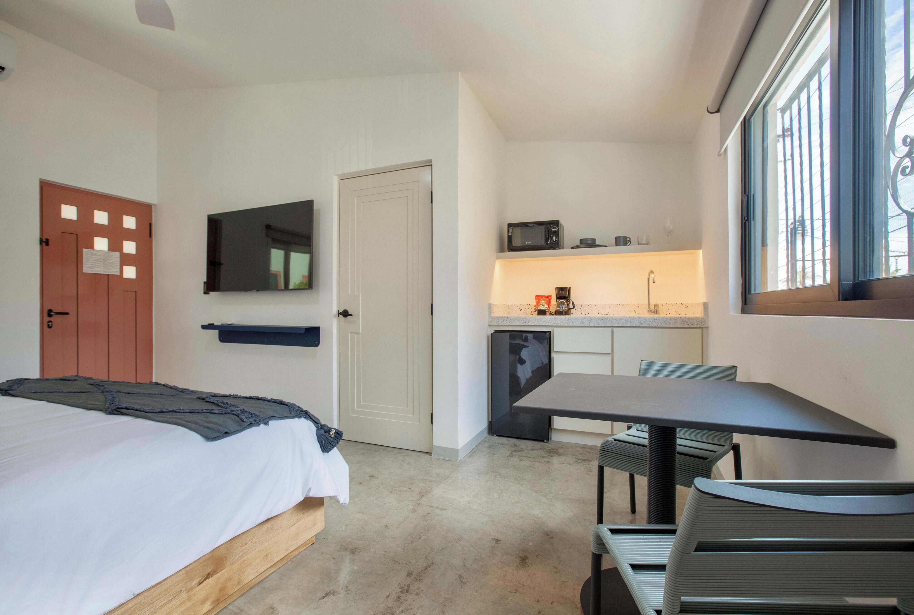 Una habitación de hotel compacta con una cama, una pequeña kitchenette, un comedor con mesa y sillas, y un televisor montado en la pared.