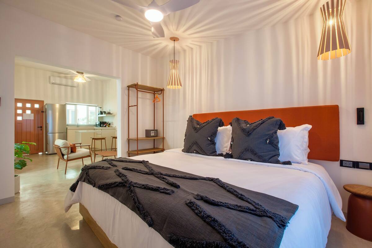 Un dormitorio moderno con una cama grande, una silla de madera, una mesita y una cocina visible a través de una puerta abierta.