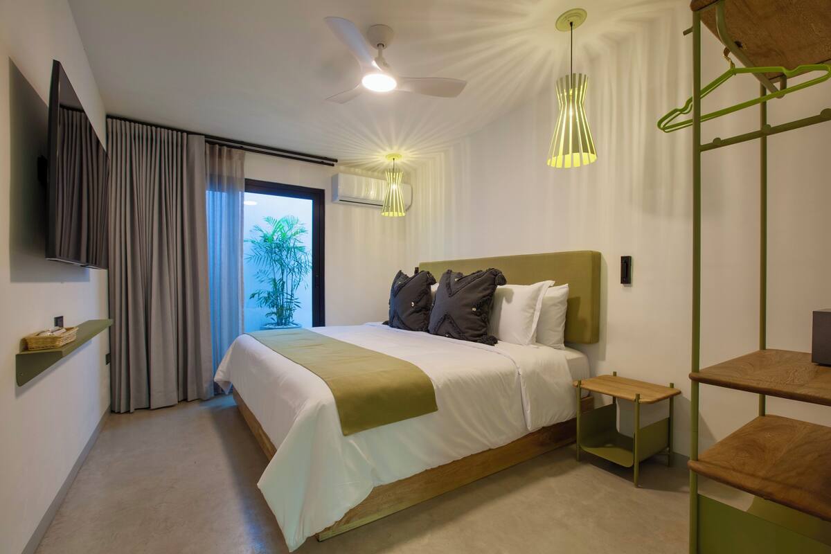 Habitación de hotel moderna con una cama grande, ventilador de techo, televisor montado en la pared y una ventana con cortinas.