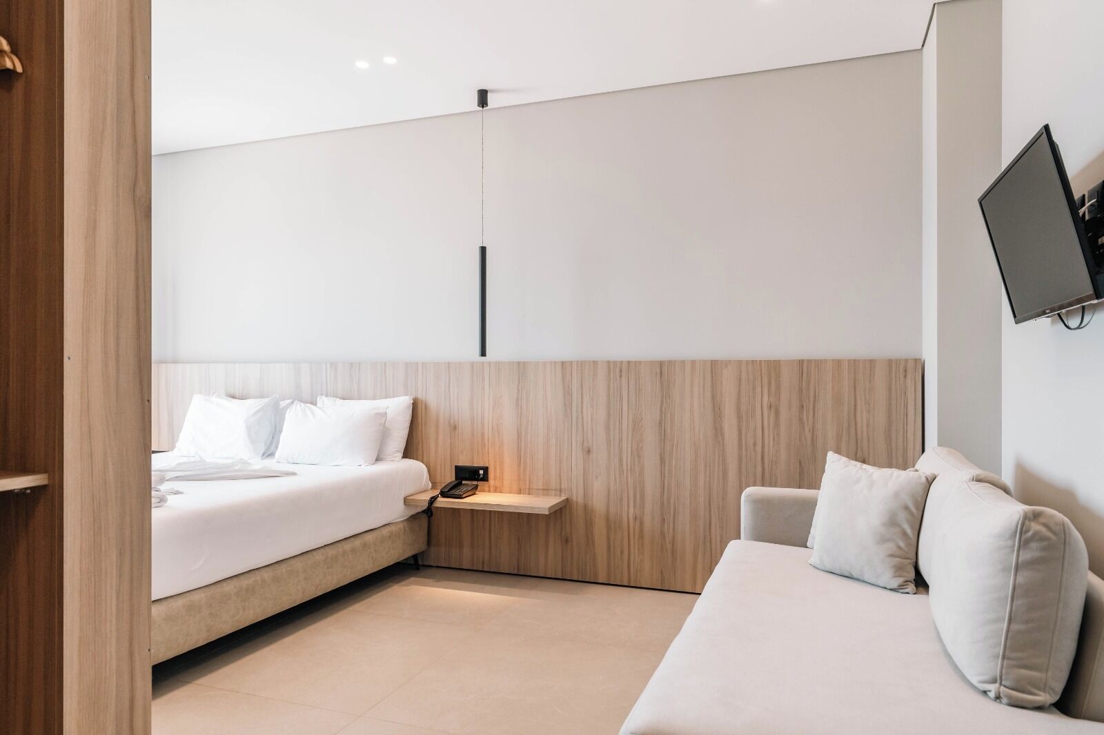 A modern hotel room with a bed, a sofa, a TV mounted on the wall, and a wooden accent wall.