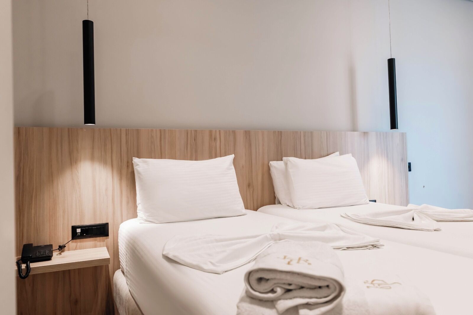 A hotel room with a wooden headboard, two white pillows, a neatly made bed with a folded towel, and a bedside table with a phone.