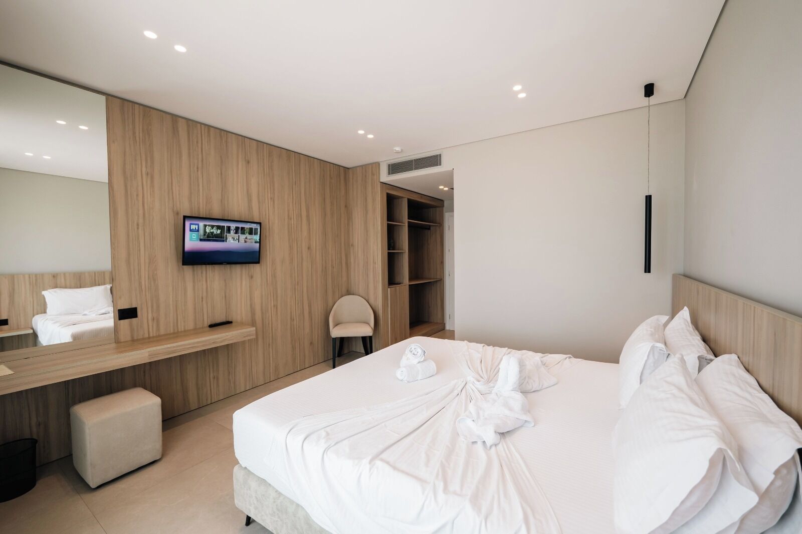 Una habitación de hotel moderna con una cama grande, un televisor en la pared, un armario de madera y una pequeña zona de estar.
