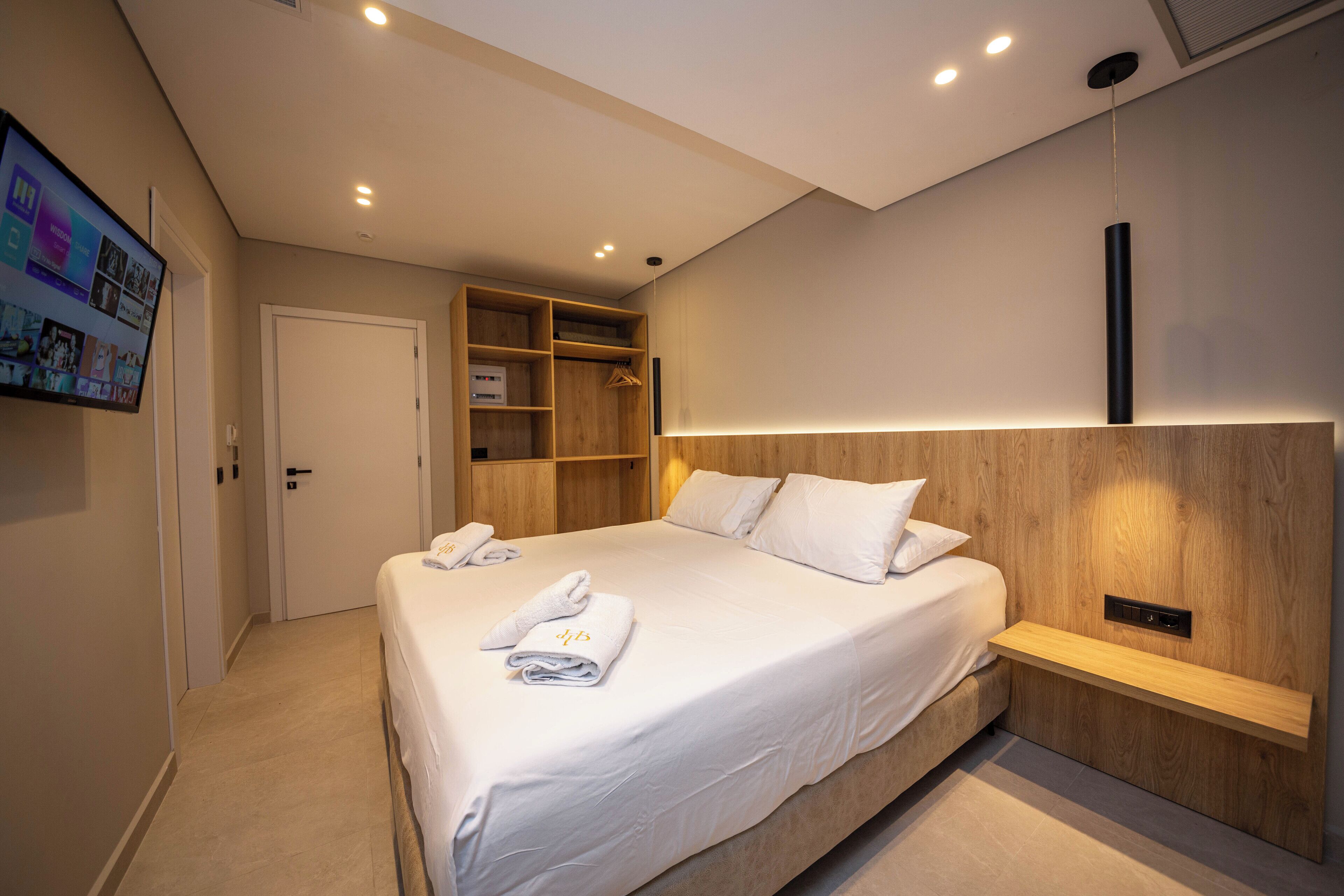 A modern hotel room with a large bed, a built-in wardrobe, a TV mounted on the wall, and a bedside table with a lamp.