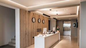 A modern reception area with a wooden accent wall, three clocks, a reception desk, and a hallway leading to an elevator. - Hotel Portanova (Sarandë)