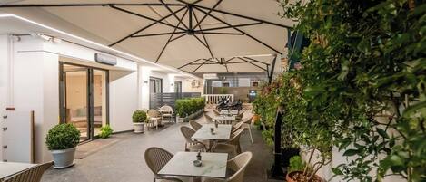 Area caffè all'aperto con tavoli, sedie e piante in vaso, con una reception sullo sfondo.