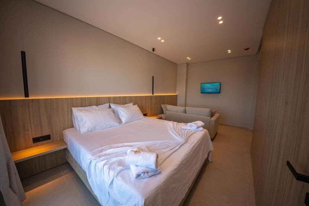 A modern hotel room with a large bed, a bedside table, a sofa, and a flat-screen TV mounted on the wall.